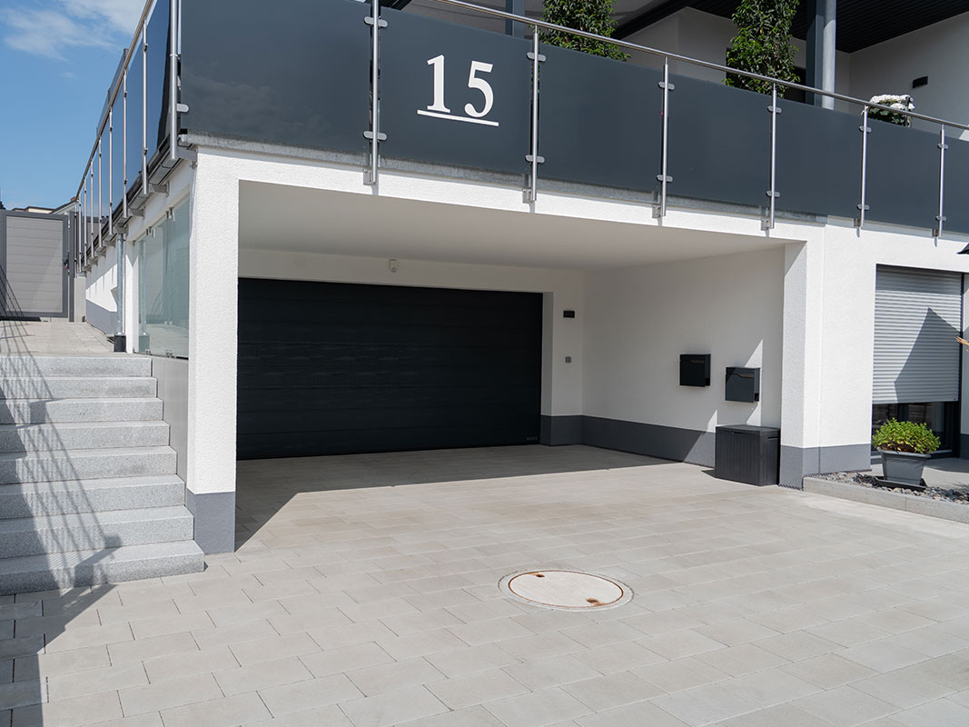Betontreppe neben großer schwarzer Garage mit gepflasterter Einfahrt in der Gartenanlage der Familie Grimm