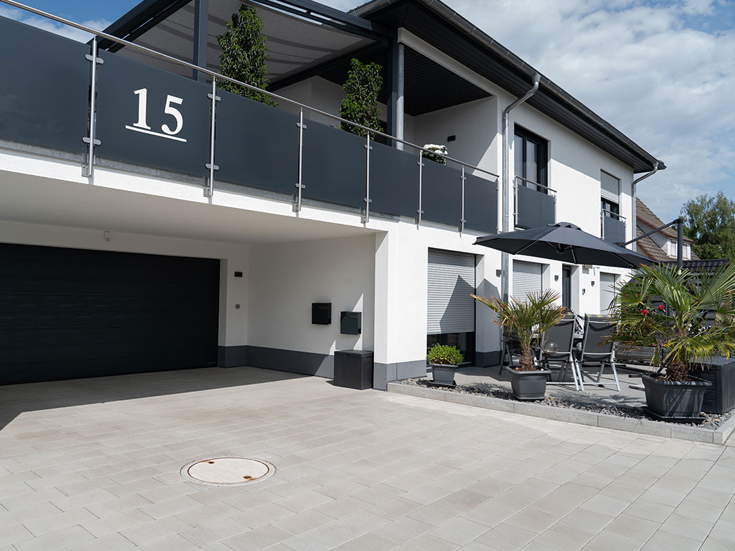 Häuserfront der Familie Grimm mit großem Garagentor; rechts davor Terrasse aus Stein mit Sonnenschirm und Sitzgruppe.