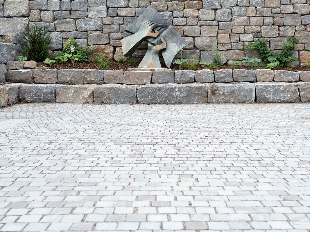Pflasterplatz mit zwei Stufen aus großen Steinen, davor eine Metallskulptur in Form eines Buches, das von zwei Händen gehalten wird, vor einer Natursteinmauer bei der Nussbaum GmbH in Rottweil.