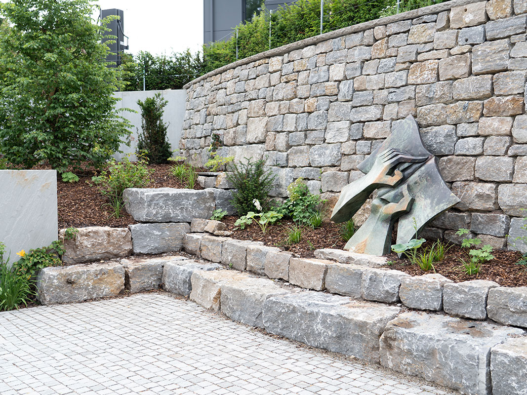 „Natursteinmauer aus unterschiedlich großen Steinen mit Torfboden, grünen Pflanzen, zwei Steinstufen und einem kupfernen Objekt.