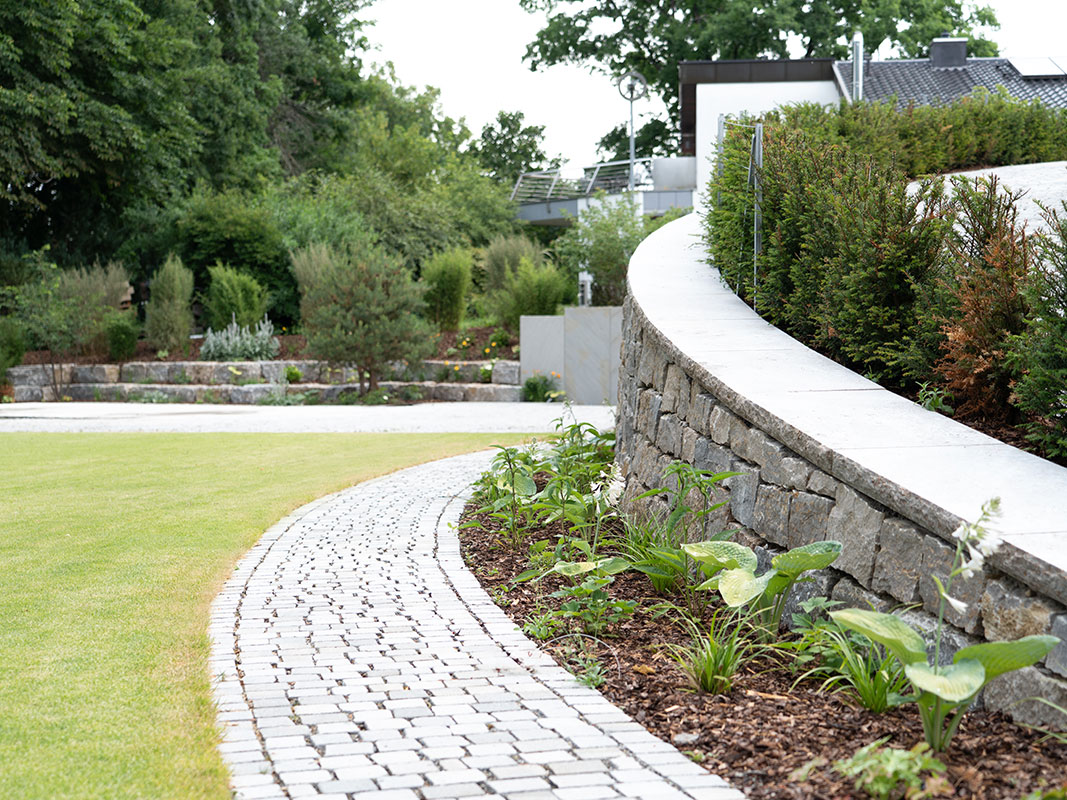 Außenanlage der Nussbaum GmbH: Rechts eine Muschelkalkmauer mit Büschen, in der Mitte ein gepflasterter Weg, rechts Rasen. Im Hintergrund stufenförmig angeordnete große Steine mit Torf und Grünpflanzen