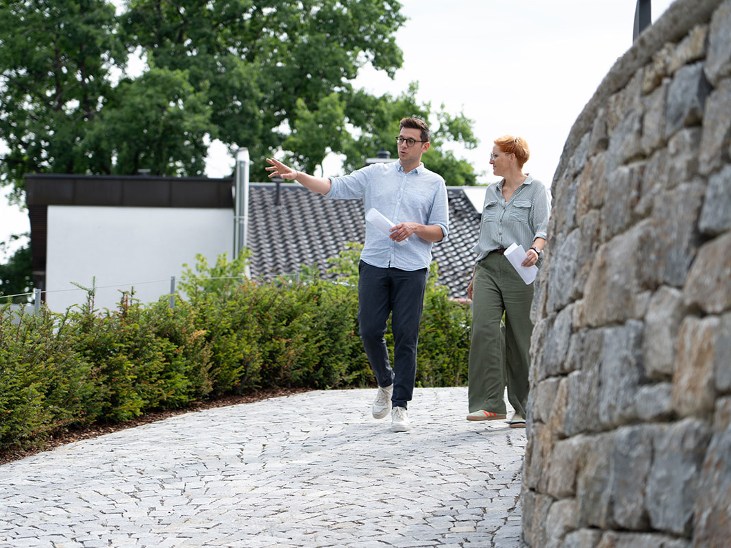 Zwei Personen gehen einen gepflasterten Weg entlang einer Natursteinmauer, rechts eine Hecke.