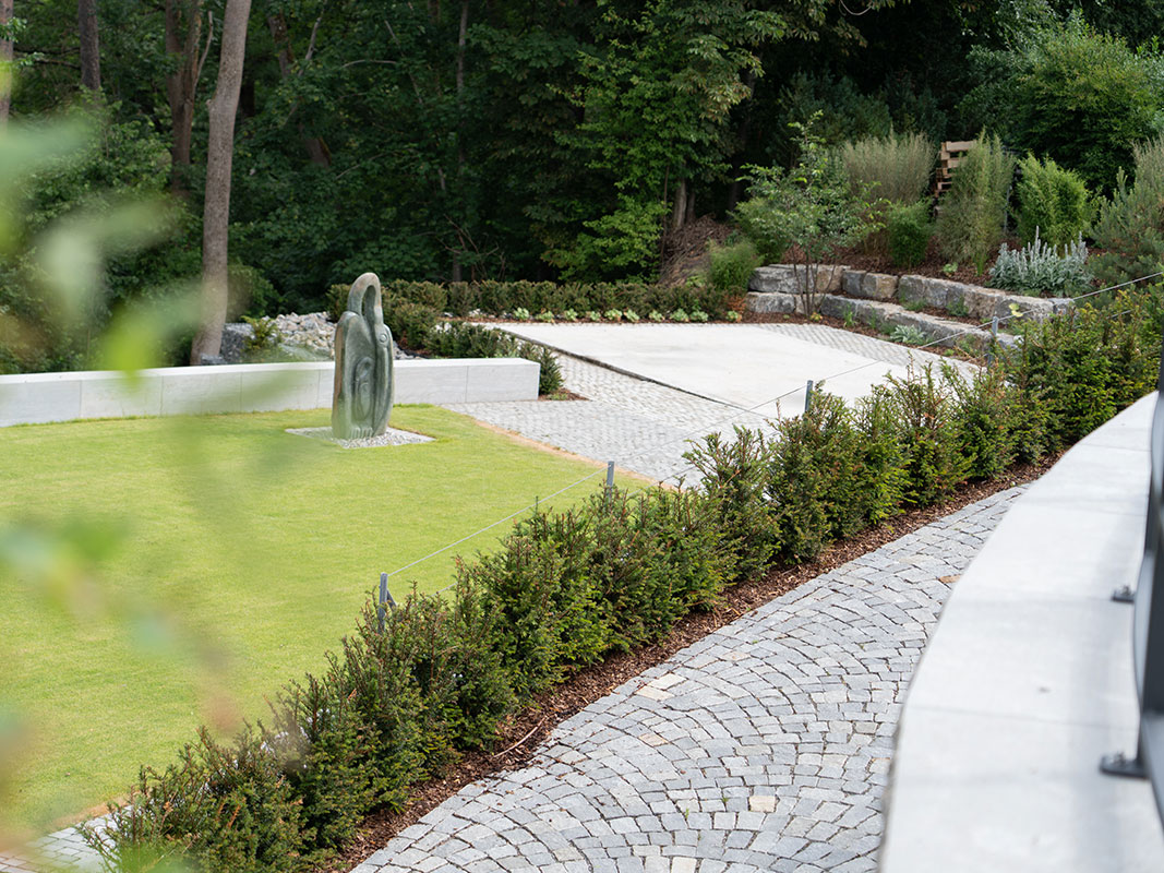 Gepflasterter Weg mit Blick auf den Garten der Nussbaum GmbH, auf dem Rasen steht eine Steinskulptur.