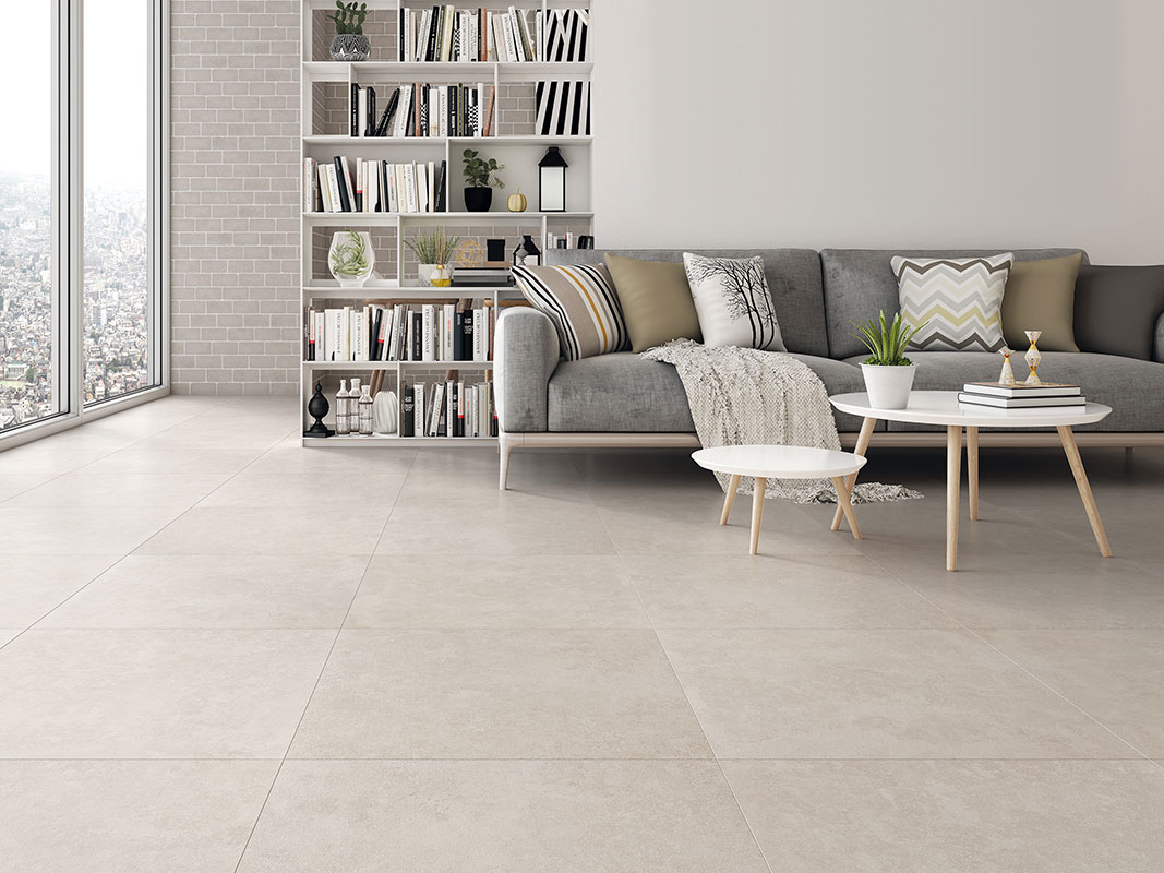 Wohnzimmer mit Fliesenboden, im Hintergrund ein weißes Regal voller Bücher. Im Vordergrund ein graues Sofa mit Kissen und ein Sofatisch.