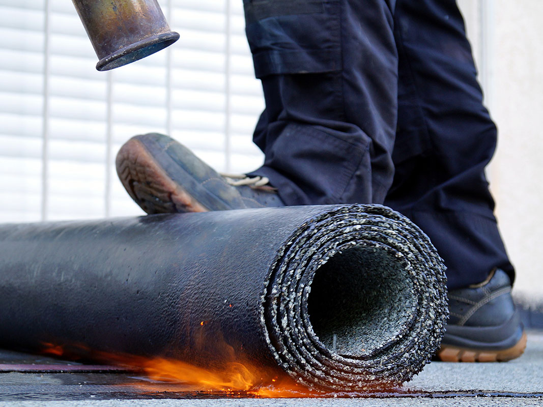 Mann beim Aufbringen von Bitumendichtungen auf einer Dachfläche, nur Füße und Bitumenrolle sichtbar.