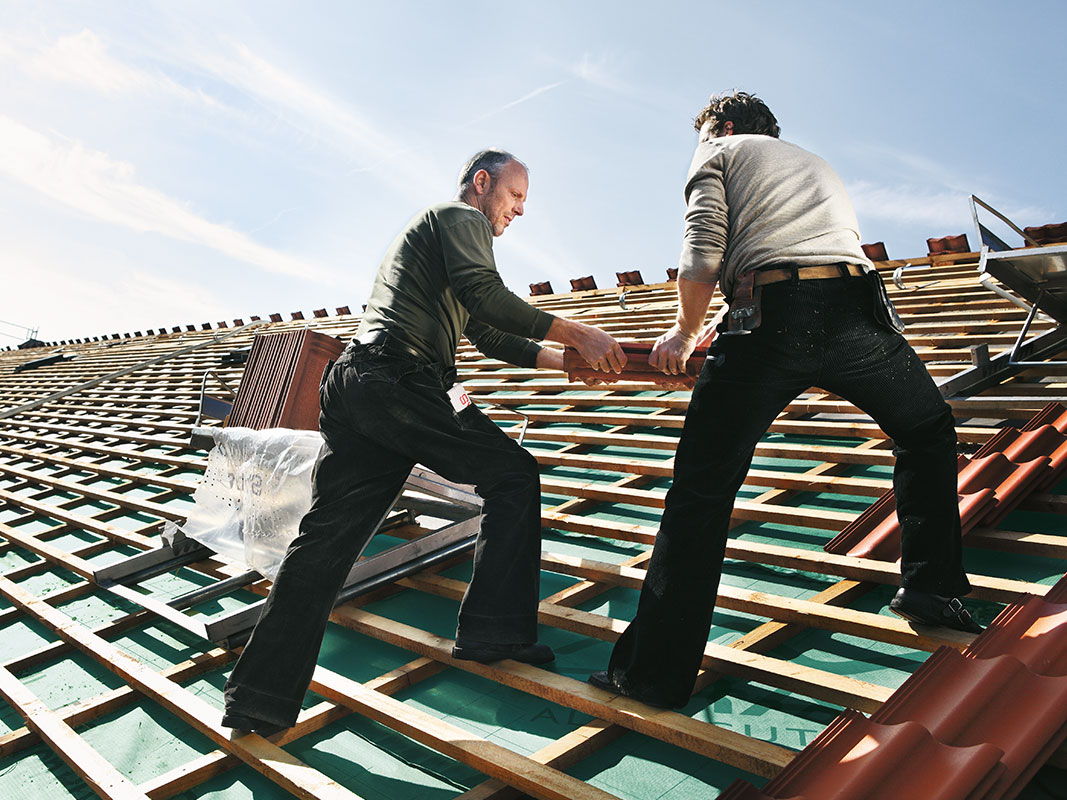 Zwei Dachdecker beim Decken eines Daches, einer übergibt dem anderen mehrere Dachziegel.