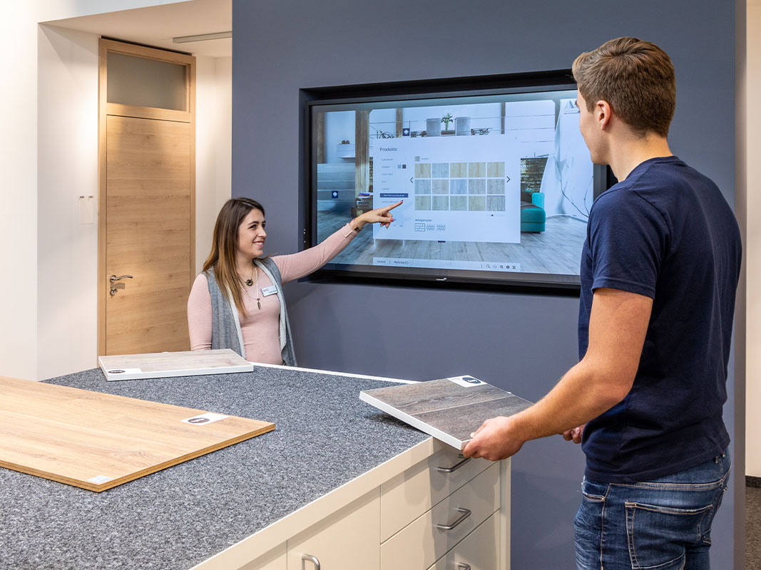 Verkäuferin mit Namensschild zeigt einem jungen Mann verschiedene Produkte auf einem in die Wand eingelassenen großen Monitor in der digitalen Ausstellung