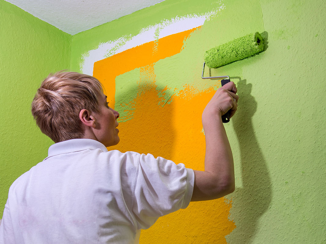Frau mit kurzen blonden Haaren und weißem Poloshirt streicht mit einer grünen Farbrolle eine orangefarbene Wand in Grün.