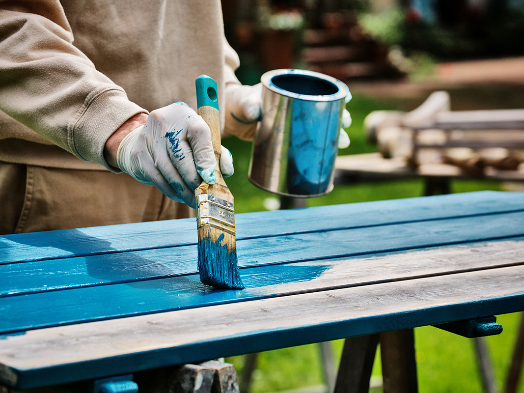 Hand mit Einweghandschuhen streicht Holzlatten mit einem Pinsel in blauer Farbe an.