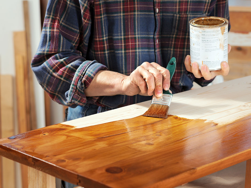 Mann im karierten Hemd trägt mit einem Pinsel eine dunkle Lasur auf eine Holzplatte auf.
