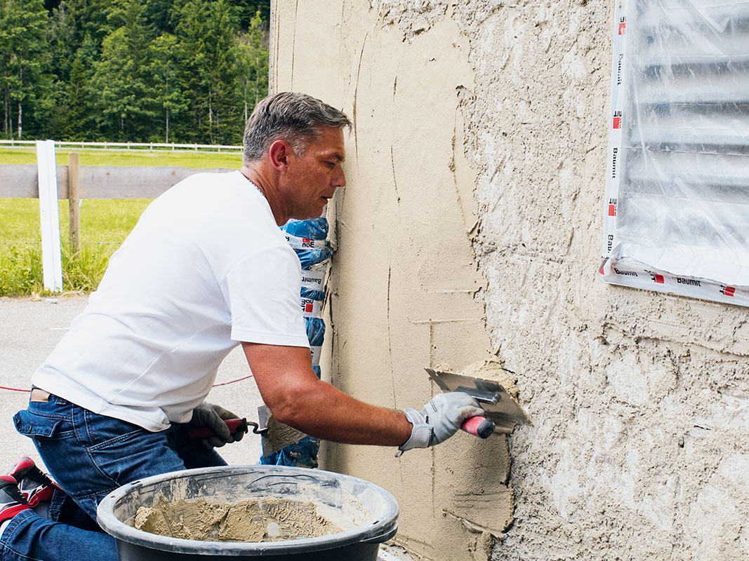 Mann trägt mit einer Glättkelle eine Putzschicht auf die Außenmauer eines Hauses auf, daneben ein großer Eimer mit Putzmasse