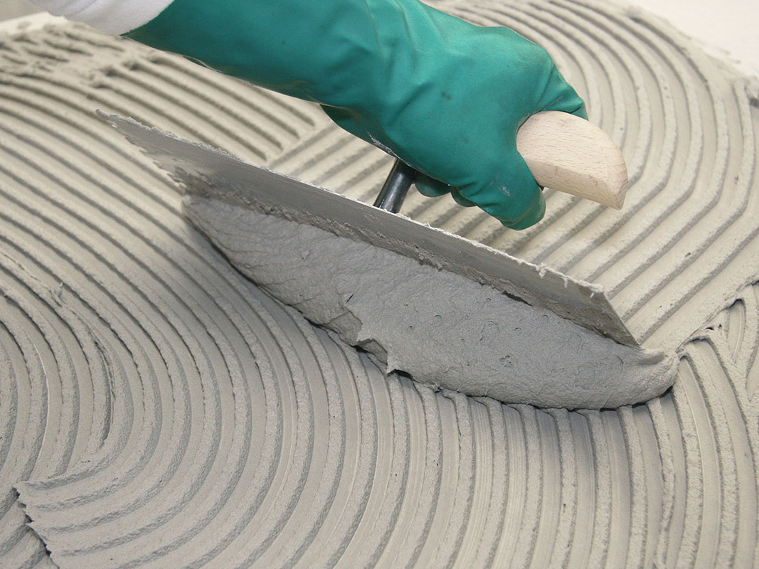 Nahaufnahme einer gezackten Glättkelle, die über Putzmasse gezogen wird; Arm mit türkisfarbenen Handschuhen.