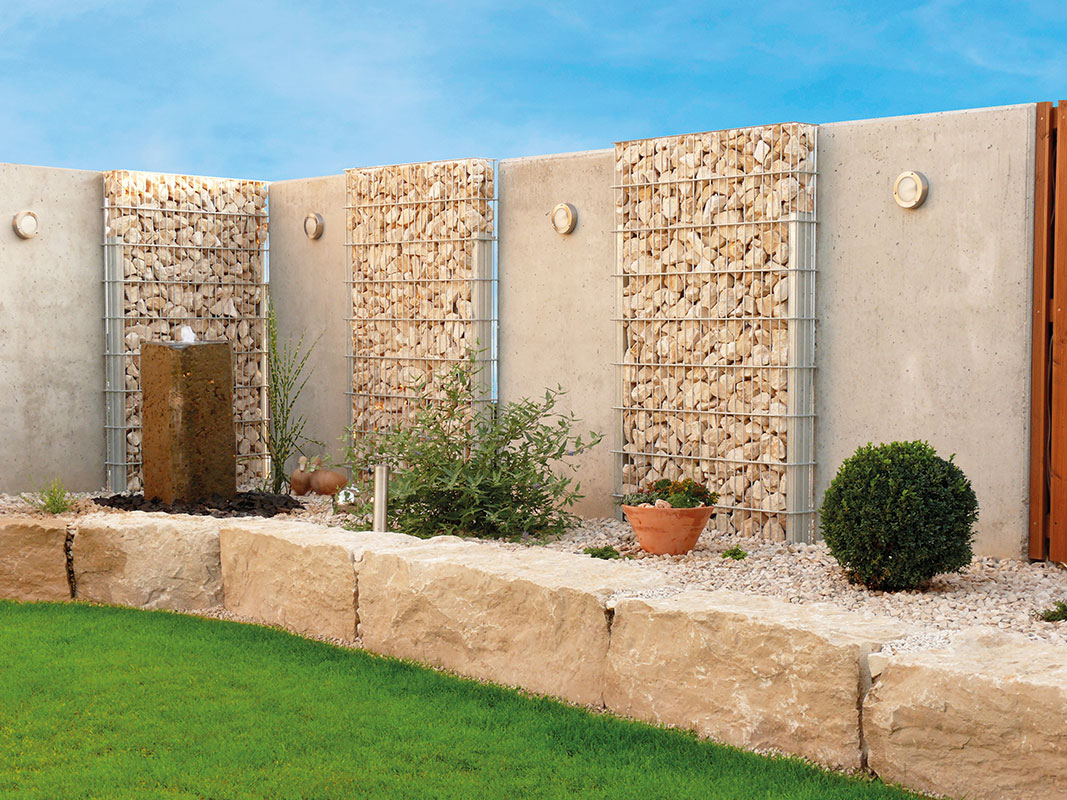 Gartenfläche mit Gabionenzäunen abgegrenzt. Vorne ein erhöhter Steingartenstreifen, getrennt vom grünen Rasen durch eine große Steinzeile.