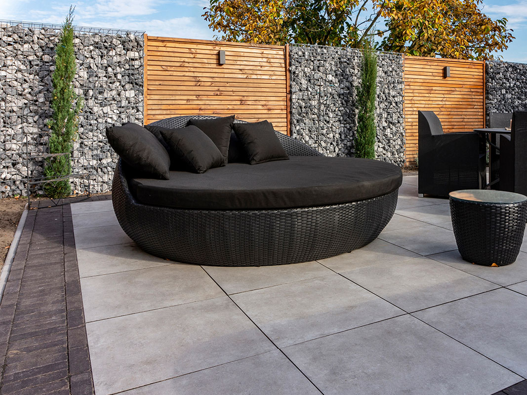 Große Terrasse mit Holz-Sichtschutz, Kiesmauern und runder schwarzer Geflecht-Liege mit Kissen.