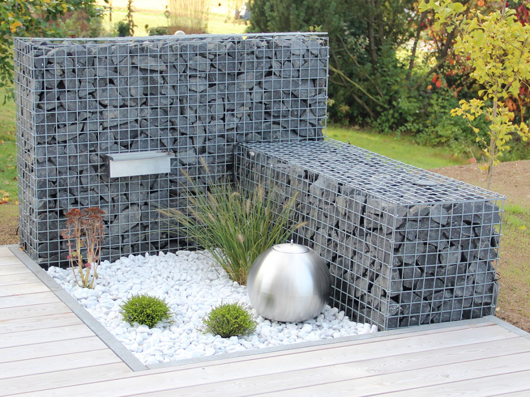 Verschiedene Gabionenkörbe und Säulen aus grauen Steinen im Garten arrangiert. Vordergrund mit weißen Steinen, Hintergrund mit grünem Garten.