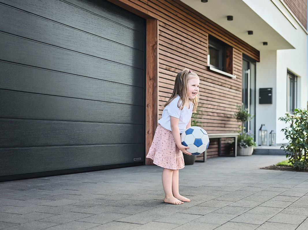 Kleines barfüßiges Mädchen hält einen Ball und steht vor einem schwarzen Garagentor im Hof; im Hintergrund ist eine Holzhauswand mit Haustür zu sehen.