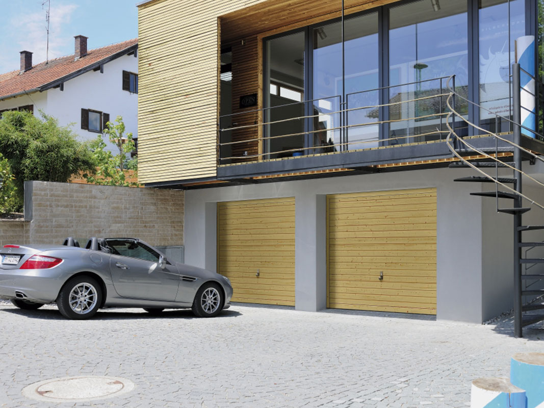 Silbernes Cabrio vor einer Doppelgarage mit Schwingtoren, darüber Hauszugang mit Wendeltreppe aus grauem Stahl und großer Glasfront.