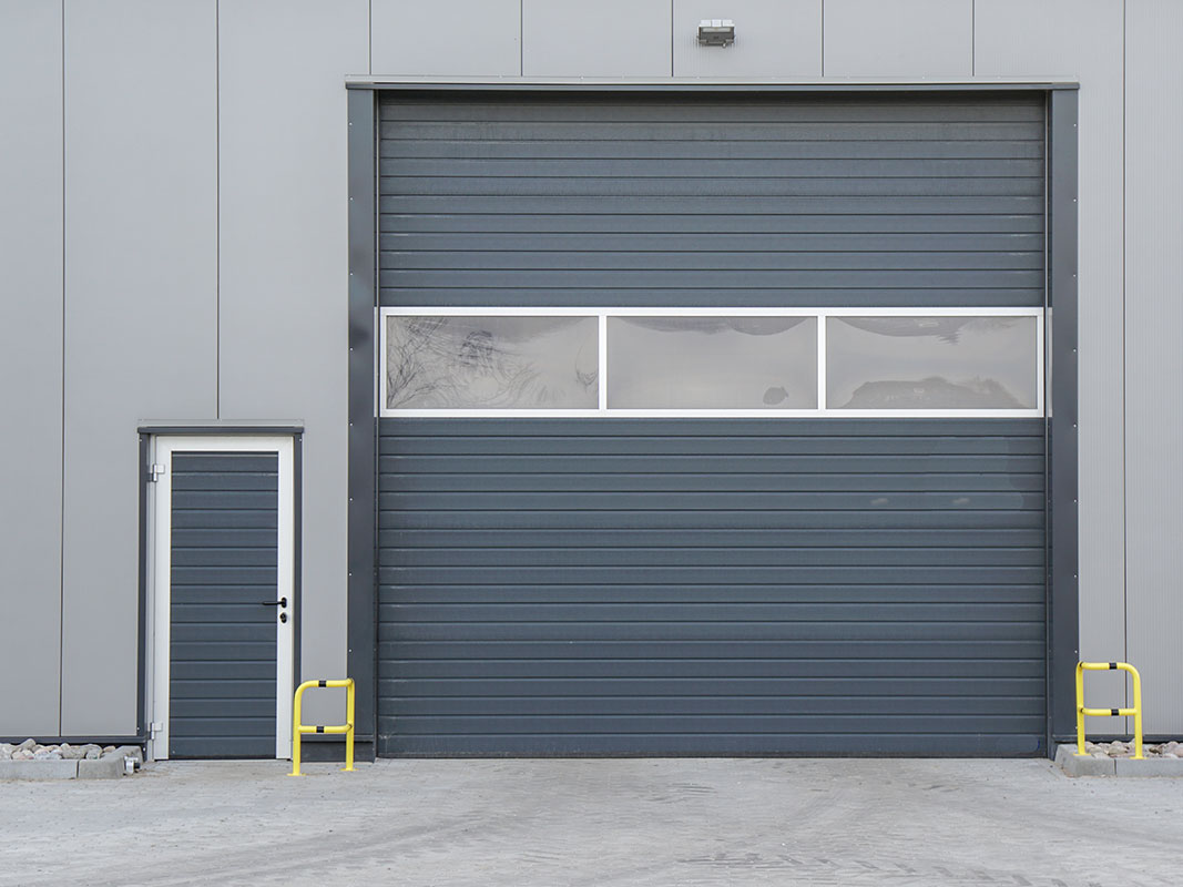 Großes dunkles Hallentor an einer grauen Halle; neben dem Tor befindet sich eine Zugangstür zur Halle.