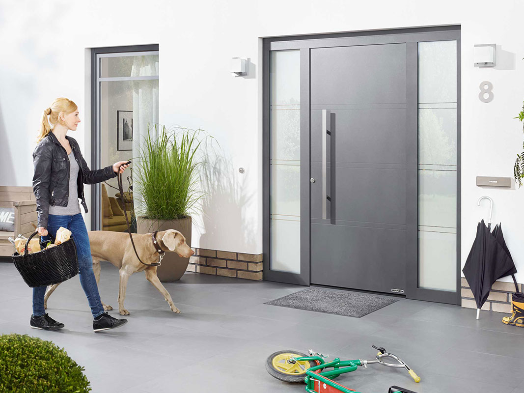 Frau mit Einkaufskorb und Hund an der Leine geht auf eine anthrazitfarbene Haustür zu; vor der Tür liegt ein Fußabstreifer, rechts davon steht ein Kinderfahrrad.