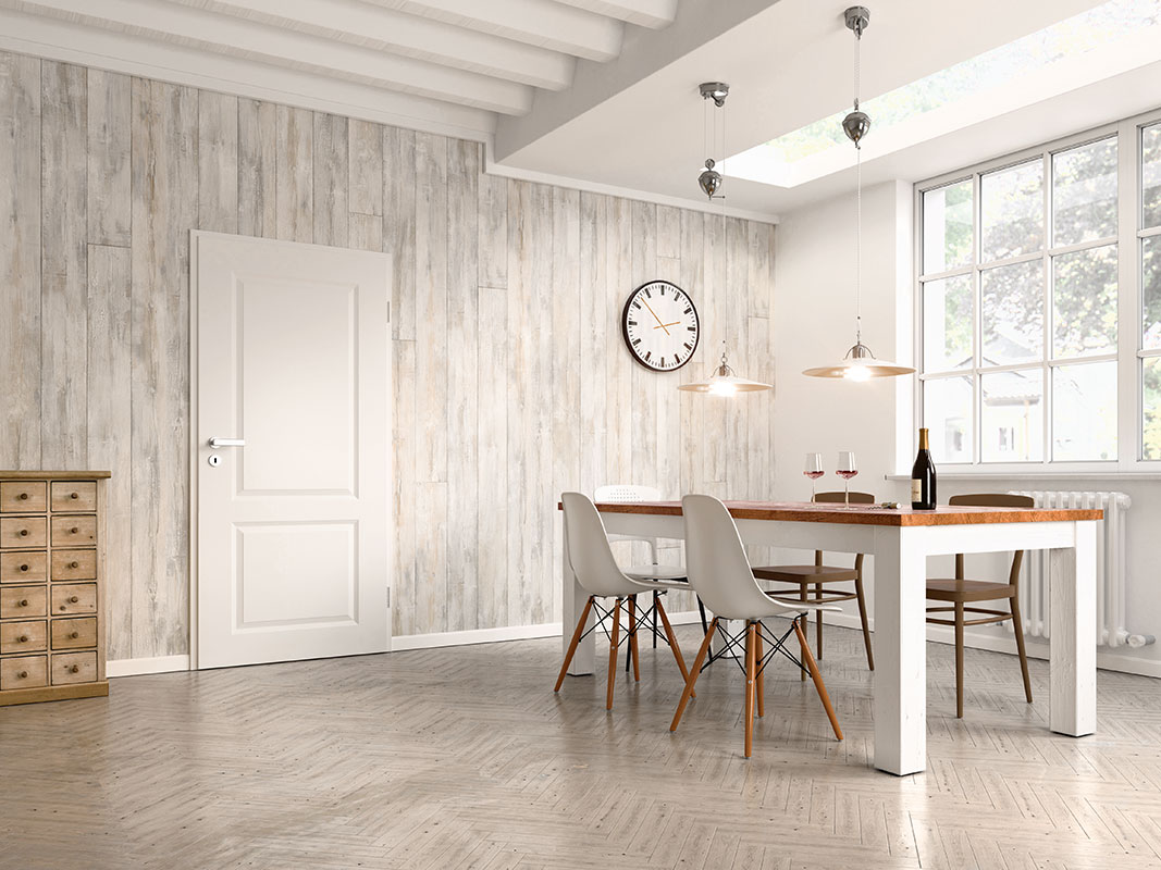 Heller Raum mit Naturholz verkleideten Wänden, weißer Tür, weißen Fensterrahmen, weißem Tisch mit Naturholzplatte und weißen Stühlen. Auf dem Tisch stehen eine Weinflasche und zwei Gläser; an der Wand hängt eine Uhr