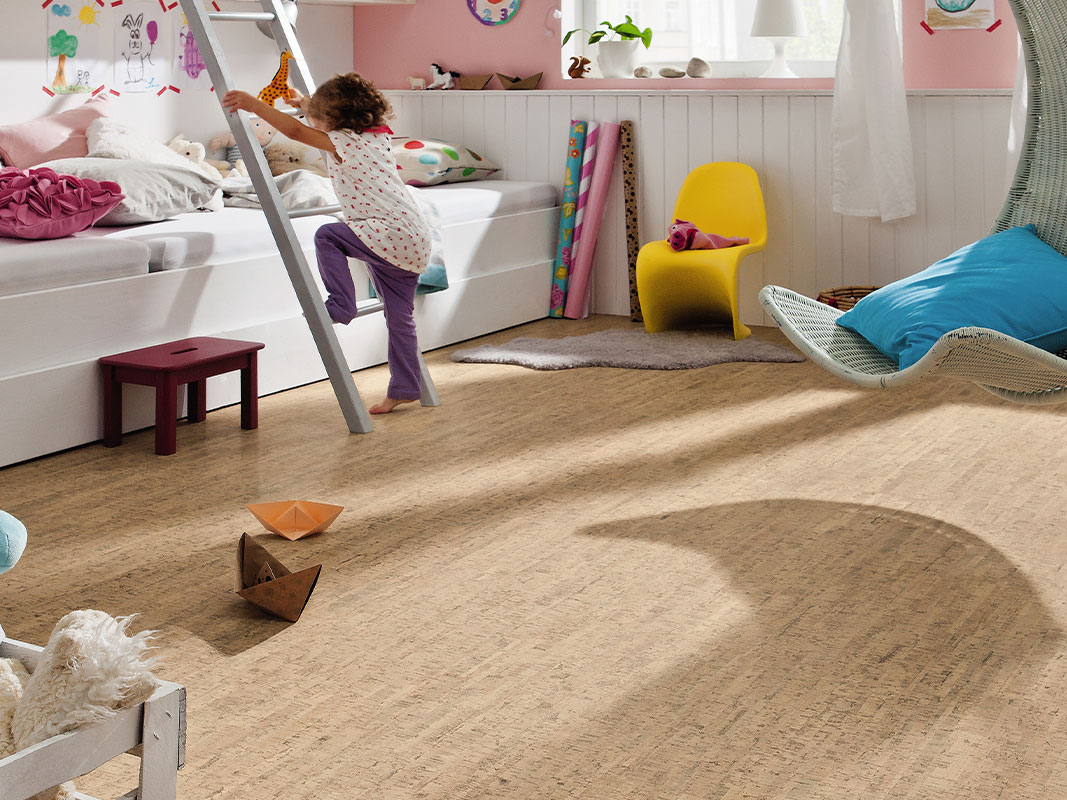 Kinderzimmer mit Korkboden, ein kleines Mädchen steigt eine Leiter hinauf, im Hintergrund ein Bett mit Kuscheltieren.