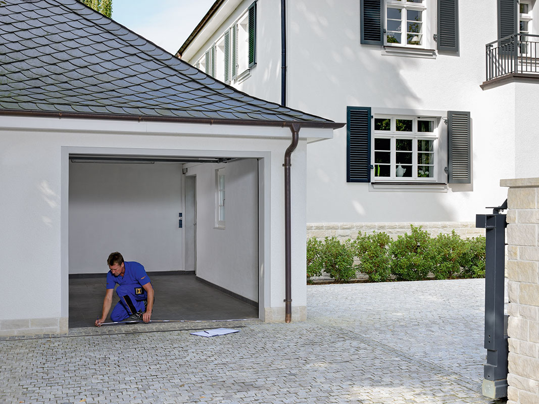Monteur misst eine Garage mit Giebeldach aus, rechts daneben ein Haus mit grauen Fensterläden.