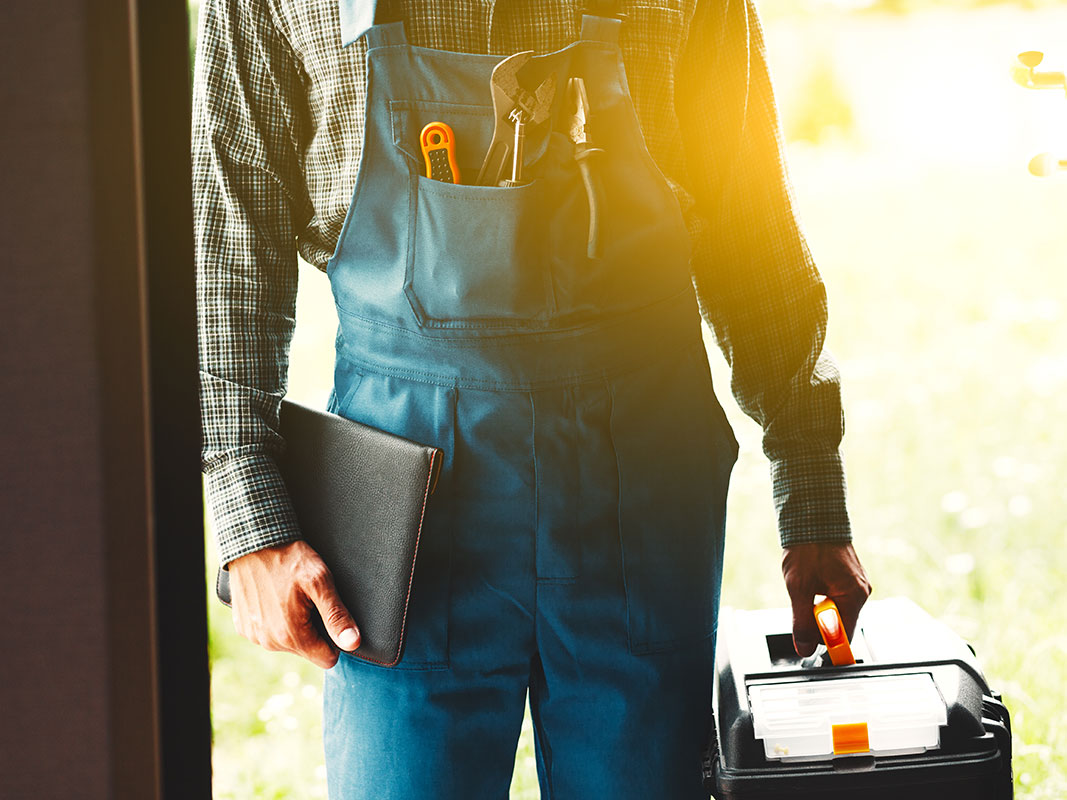 Mann im Blaumann hält in der einen Hand eine schwarze Mappe und in der anderen einen Werkzeugkoffer, bereit für Montage- und Ausbauarbeiten.
