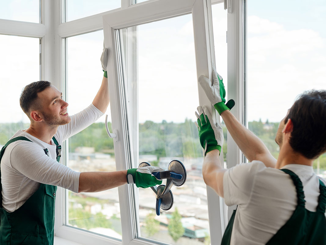 Zwei Männer in grünen Overalls setzen ein weiß gerahmtes Fenster fachgerecht ein.