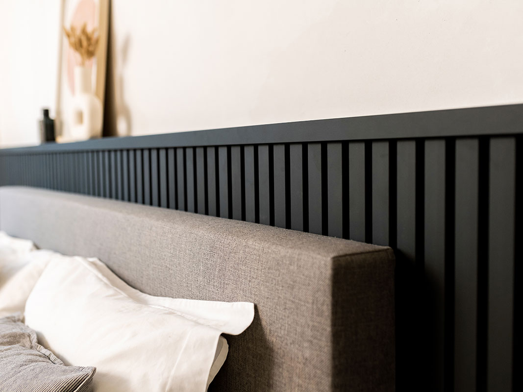 Nahaufnahme einer stoffbezogenen Bettkante, dahinter moderne schwarze Wandpaneele als stilvolles Gestaltungselement.