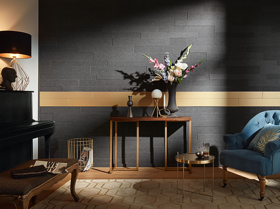 Wohnraum mit schwarzer Dekorpaneele mit goldener Linie, davor kleiner Tisch mit schwarzer Blumenvase, blauer Samtsessel, goldener Papierkorb und angeschnittenes schwarzes Klavier.