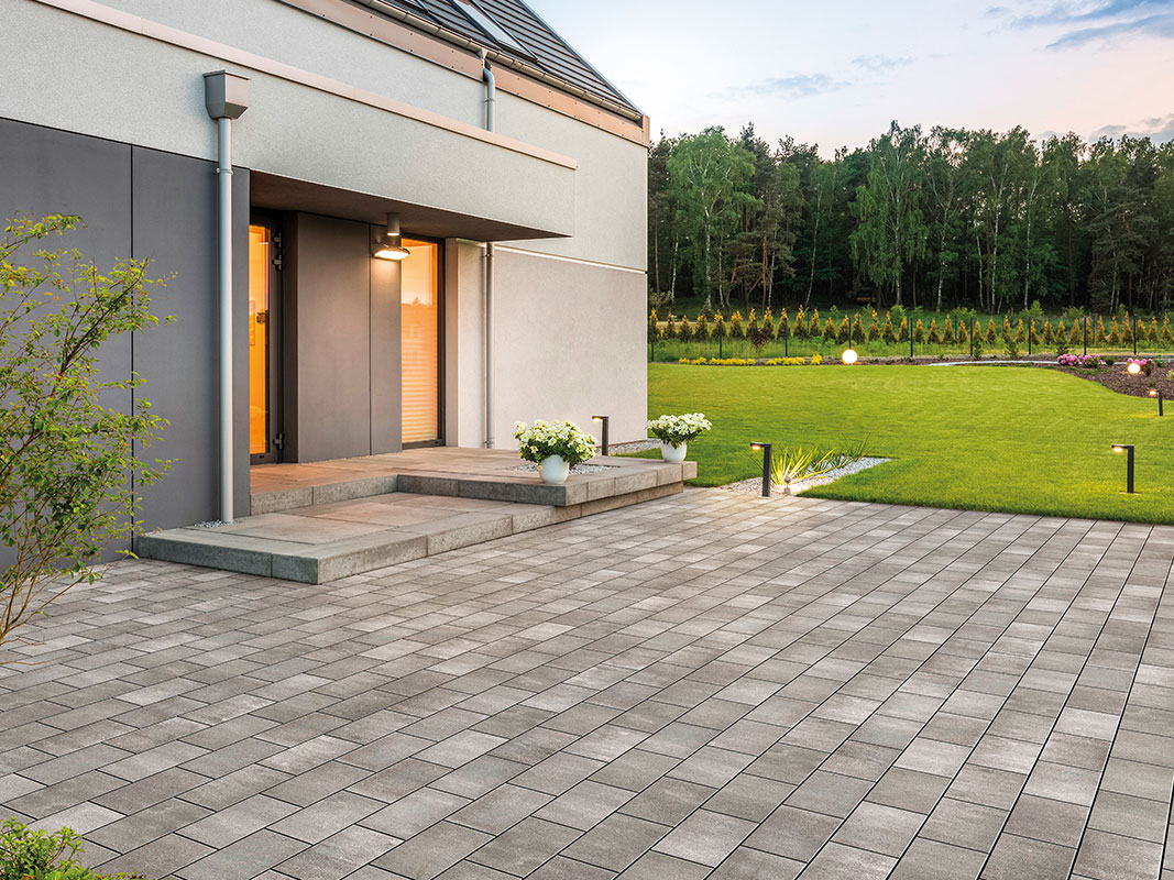 Betonpflastersteine auf einem Hof verlegt; rechts davon grüner Rasen, links ein Haus mit beleuchtetem Eingangsbereich.