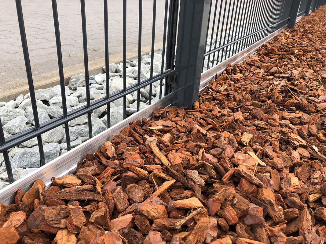 Aufgeschütteter Rindenmulch neben einem Metallzaun, im Hintergrund Steine und ein gepflasterter Weg, als Beispiel für dekorative Schüttgüter im Garten.