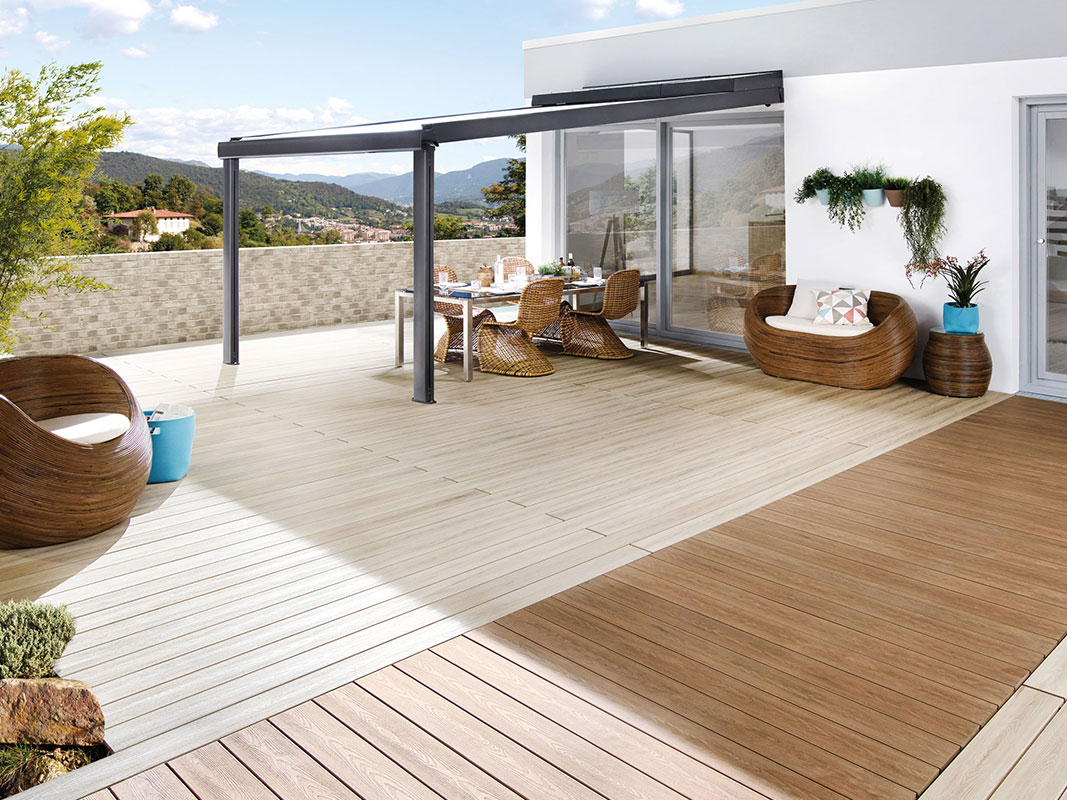 Terrasse mit verschiedenen Holzbodenbereichen und unterschiedlichen Sitzgelegenheiten aus Holz. Ein Bereich ist mit Glas überdacht; darunter steht ein gedeckter Tisch mit geflochtenen Holzstühlen.