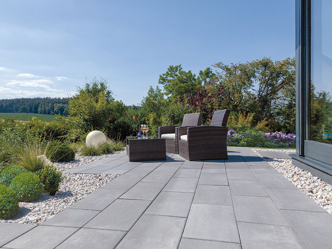 Terrasse aus Betonwerksteinplatten, umrandet mit weißen Kieseln. Im Vordergrund stehen zwei dunkel geflochtene Gartensessel mit Tisch; im Hintergrund ein grüner Garten.