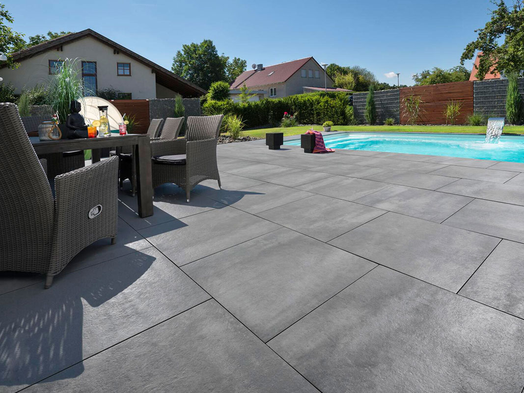 Terrasse mit dunklen Terrassenplatten, im Hintergrund ein Pool. Im Vordergrund steht ein dunkler Holztisch mit passenden, geflochtenen Stühlen.