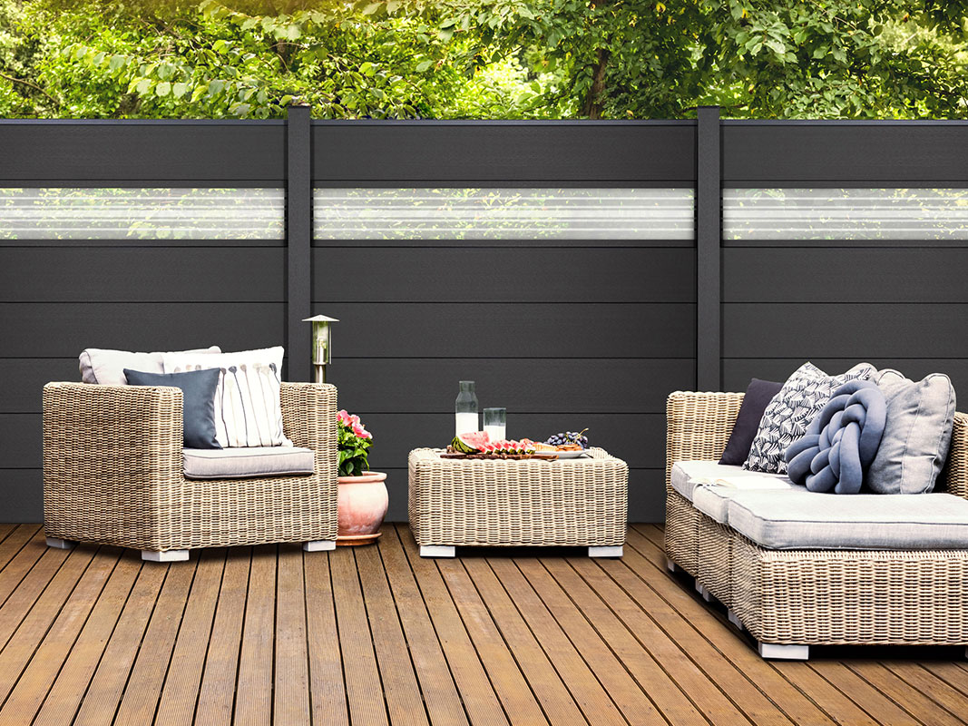 Holzterrasse mit schwarzem Sichtschutz, im Vordergrund eine geflochtene Sitzgruppe bestehend aus Sessel, Tisch und Liege, ausgestattet mit vielen Kissen.