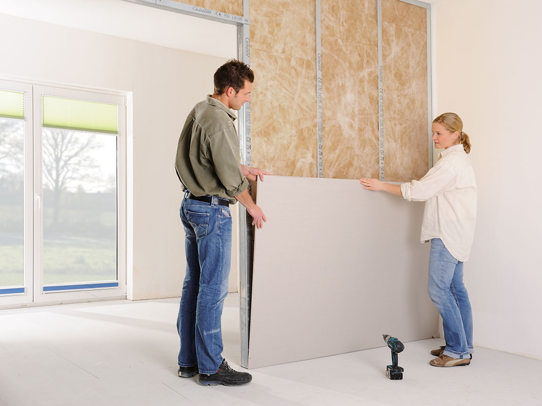 Ein Mann und eine Frau passen gemeinsam eine Gipsplatte an eine Wand an – Beispiel für Trockenbau-Arbeiten.