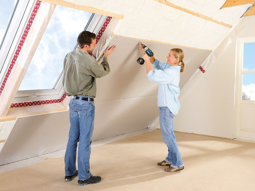 Mann und Frau bringen gemeinsam Trockenbauplatten an einer Dachschräge an; er hält die Platte, sie schraubt sie mit einem Elektroschrauber fest.
