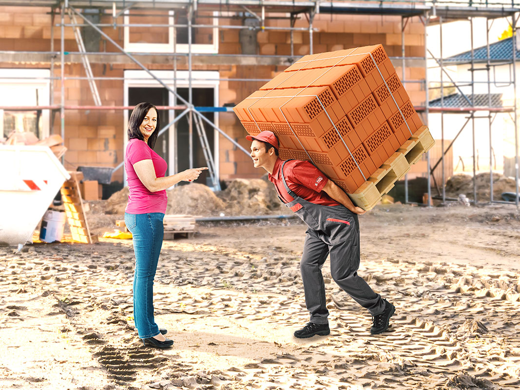 Rohbau auf einer Baustelle: Eine Frau zeigt auf einen Mann, der eine Palette voller Mauerziegel auf dem Rücken trägt.