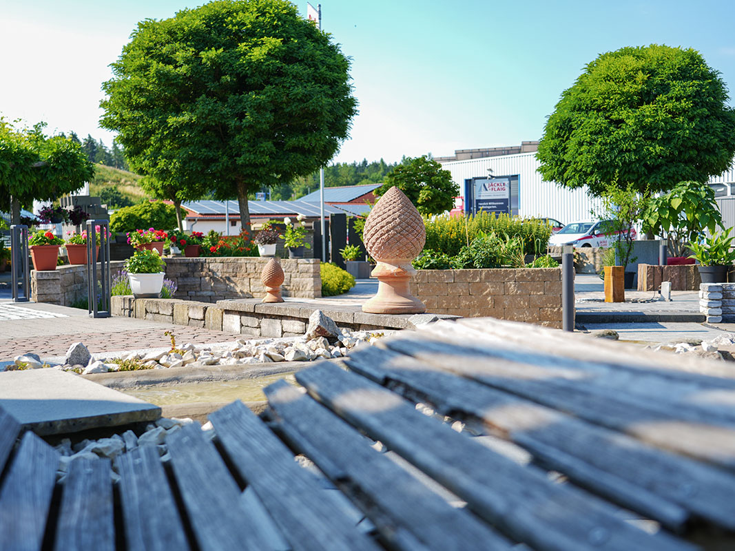 Steingartenausstellung in Niedereschach mit Holzliege im Vordergrund und Gartenskulpturen im Hintergrund.