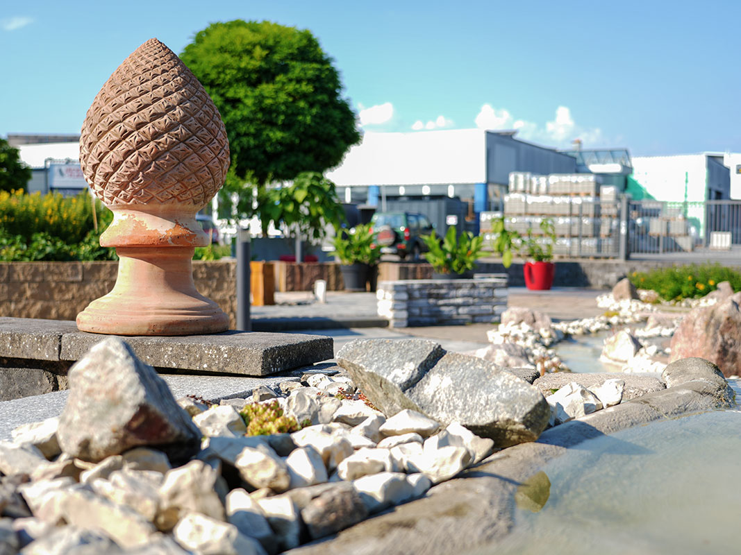 Steingartenausstellung in Niedereschach mit kleinem Bachlauf und Terrakotta-Gartenskulptur in Form eines Tannenzapfs, im Hintergrund das Gebäude von Jäckle+Flaig