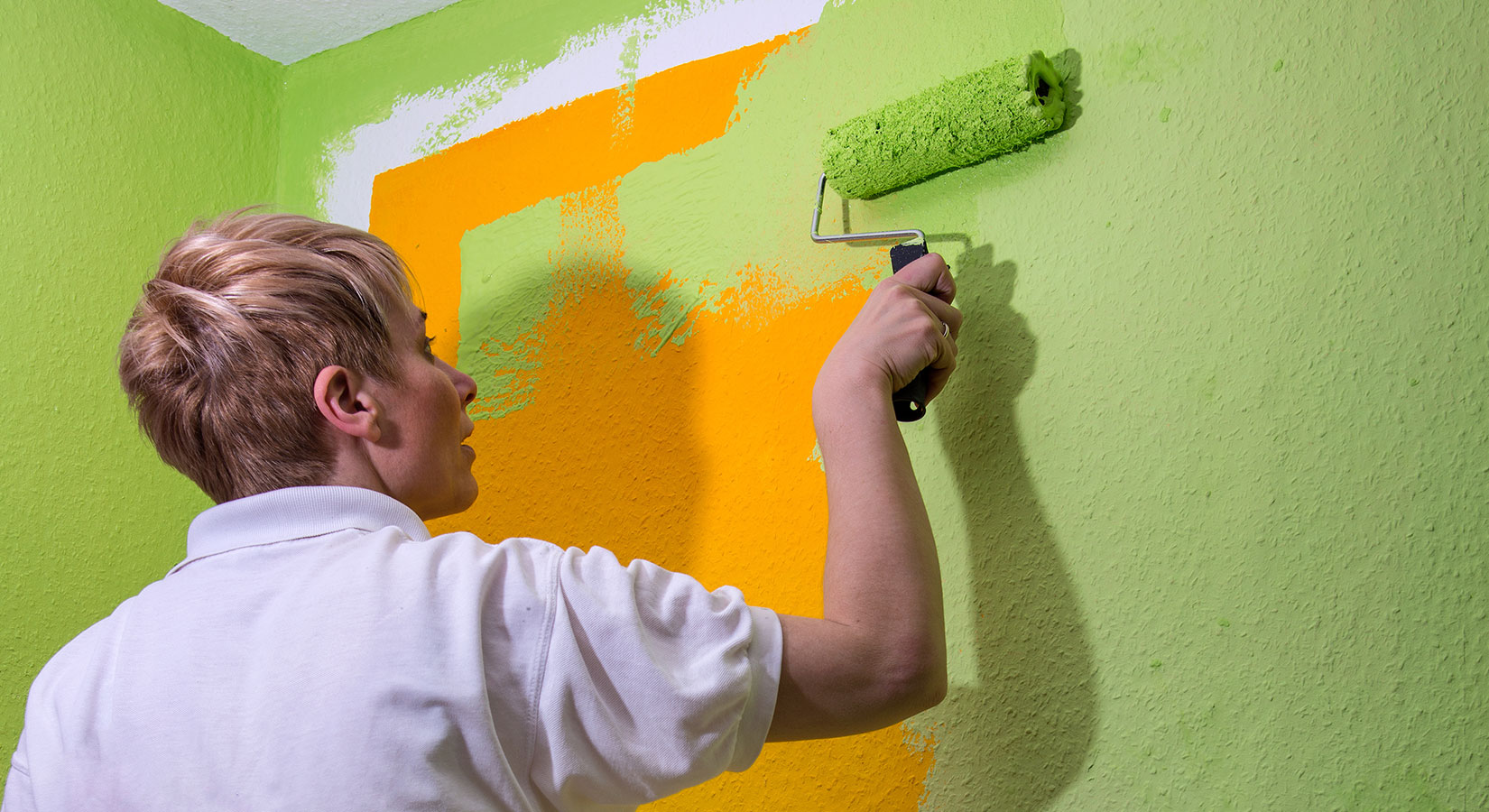 Frau mit kurzen blonden Haaren und weißem Poloshirt streicht mit einer grünen Farbrolle eine orangefarbene Wand in Grün.