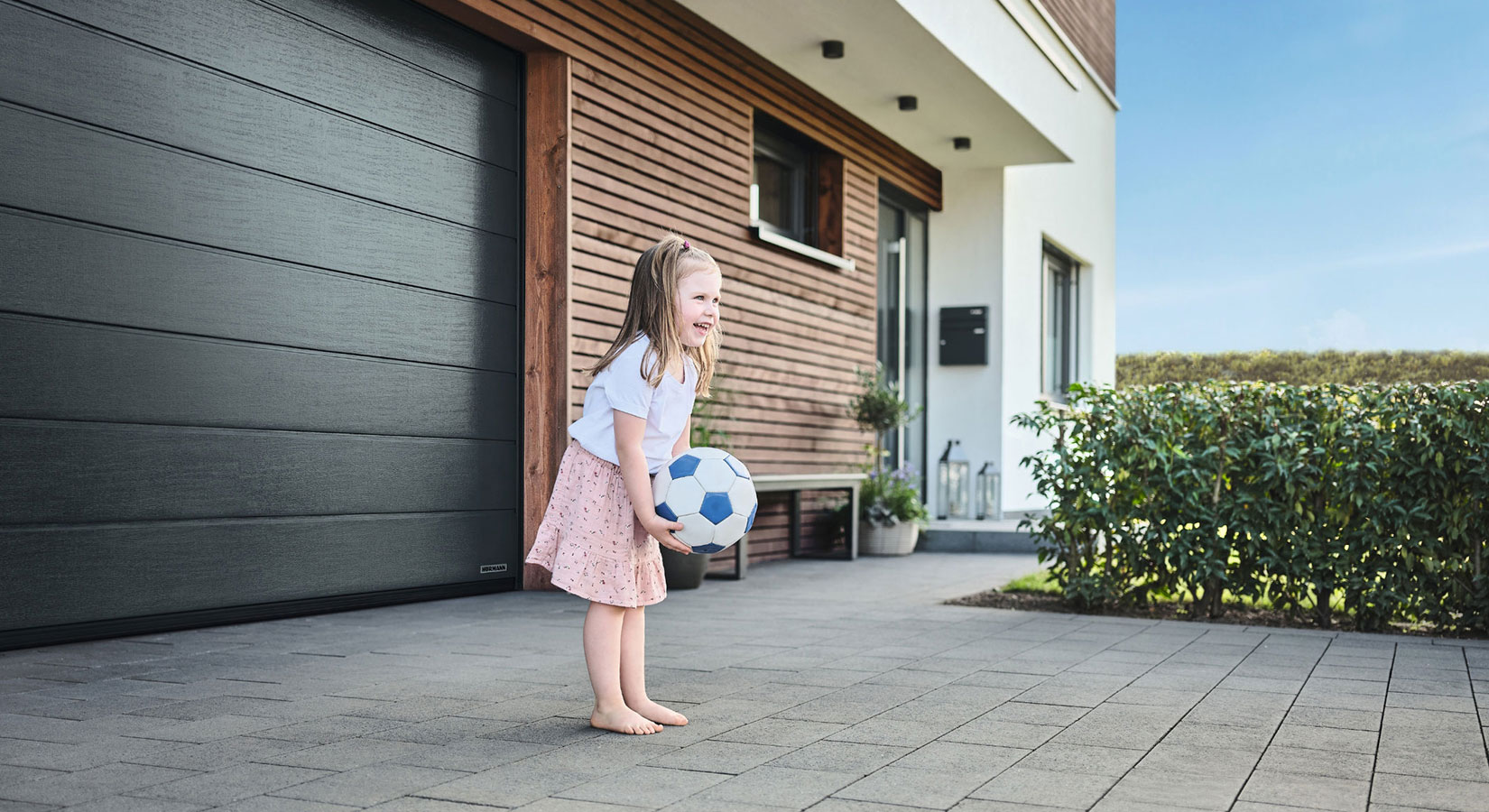 Kleines barfüßiges Mädchen hält einen Ball und steht vor einem schwarzen Garagentor im Hof; im Hintergrund ist eine Holzhauswand mit Haustür zu sehen.