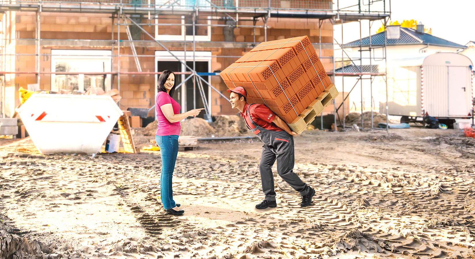 Rohbau auf einer Baustelle: Eine Frau zeigt auf einen Mann, der eine Palette voller Mauerziegel auf dem Rücken trägt.