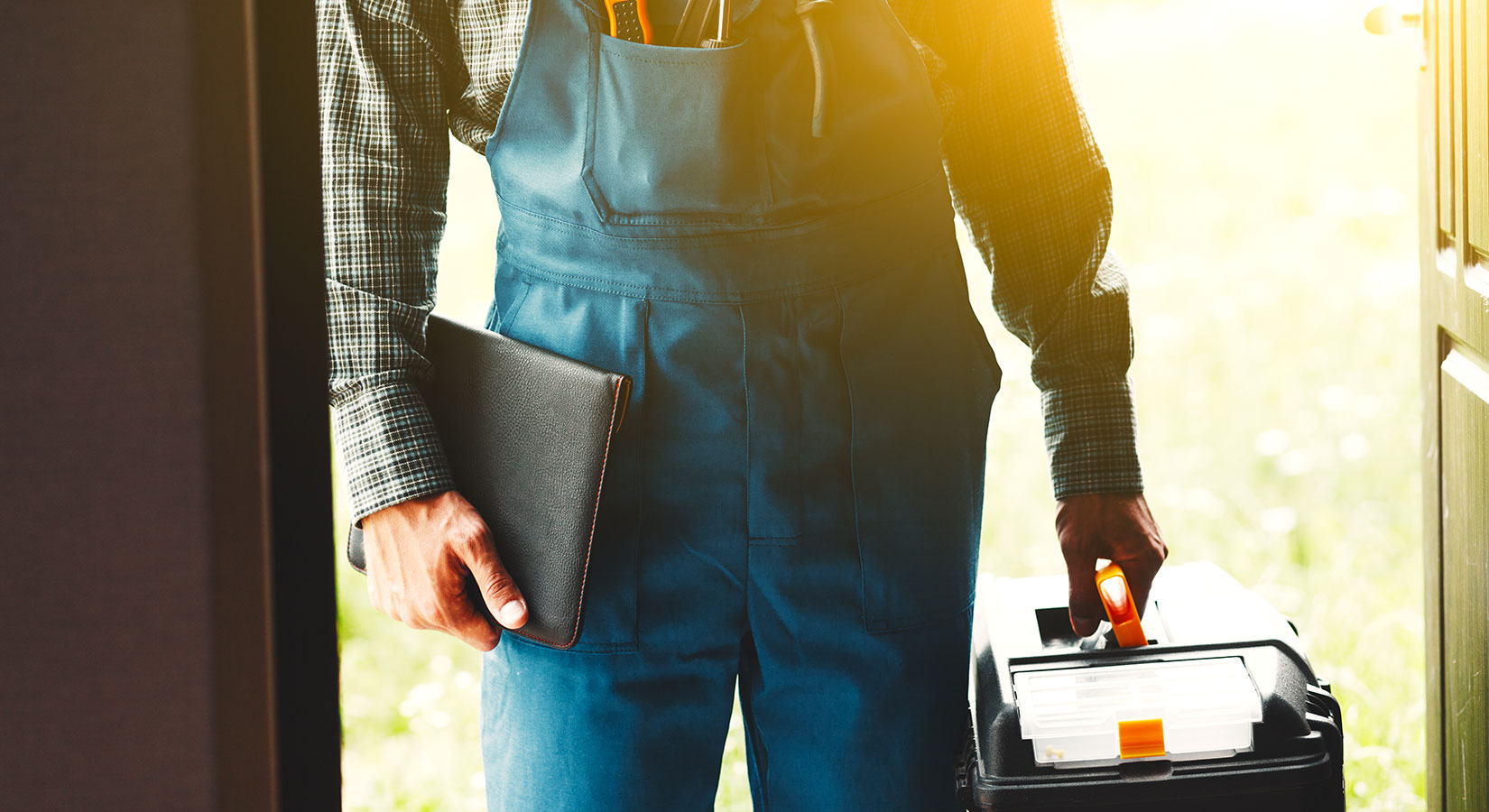 Mann im Blaumann hält eine schwarze Mappe in der einen Hand und einen Werkzeugkoffer in der anderen Hand.