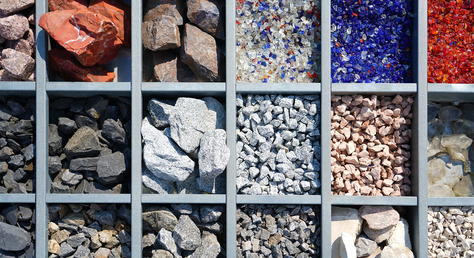 Von oben fotografierte Schublade mit mehreren Fächern, die verschiedene Steinsorten enthalten – von großen schwarzen Steinen bis zu blauen und roten Glassteinen.