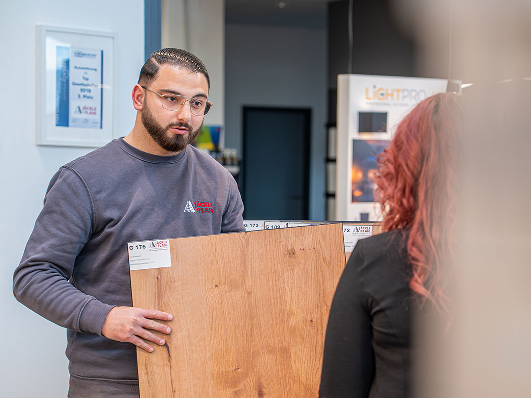 Verkäufer in grauem Jäckle+Flaig-Pullover zeigt einer Kundin verschiedene Bodenbeläge